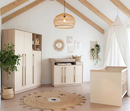 Ein helles Kinderzimmer mit einem Roba 3-teiligen Kinderzimmer-Set »Elva« in Cashmere und Virginia Chestnut, bestehend aus einem Kleiderschrank, einer Wickelkommode und einem Babybett. Ein runder Bären-Teppich liegt auf dem Holzboden.