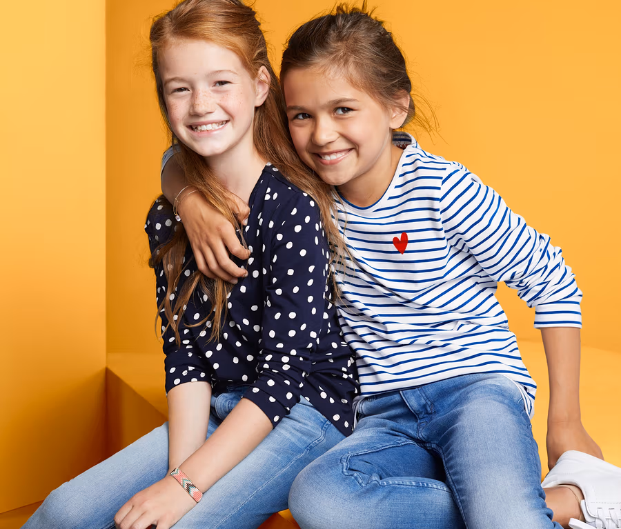Mädchen sitzen in 2 Langarmshirts. Ein Mädchen trägt ein gepunktetes Shirt, das andere ein gestreiftes Shirt.