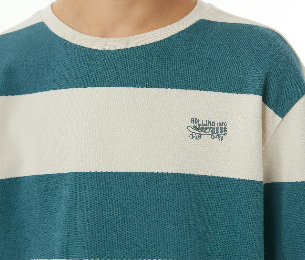 Nahaufnahme einer Person in einem gestreiften SCHIESSER Kinder-Interlock-Pyjama-Oberteil. Es hat cremefarbene und petrolfarbene Streifen sowie eine Skateboard-Stickerei mit 'ROLLING INTO HAPPINESS'.