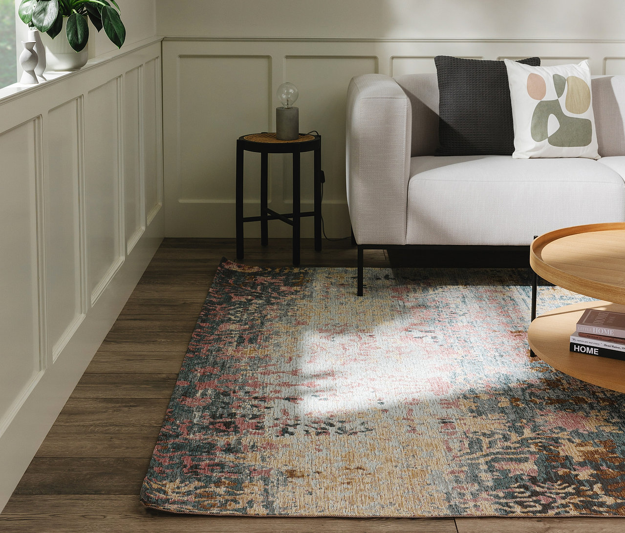 Wohnzimmerecke mit hellgrauem Sofa, Holztisch und einem Benuta Teppich »Frencie« auf Holzfußboden. Der Teppich zeigt ein abstraktes Muster in Rosa, Blau, Gelb und Beige.
