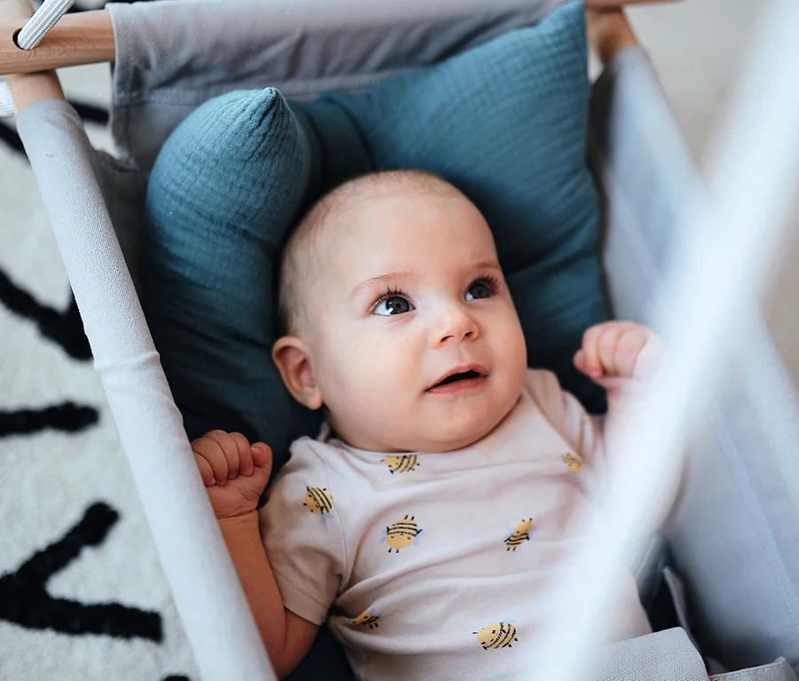 Ein Baby liegt in einer grauen small foot Babyschaukel »Seaside« mit blauem Kissen auf einer Unterlage mit schwarz-weißem Alphabet-Muster.