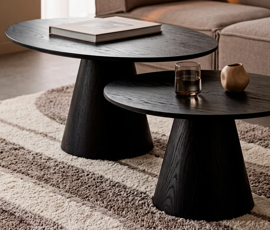 Zwei schwarze, runde WOOOD Couchtische »Posture« (ca. 120 cm und 70 cm Durchmesser) stehen ineinander verschachtelt auf einem gestreiften Teppich. Auf dem größeren Tisch liegt ein Buch, auf dem kleineren ein Glas Wasser und ein Deko-Objekt. Im Hintergrund ist ein beiges Sofa zu sehen.