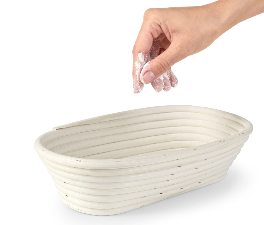 Eine Hand streut Mehl in ein ovales Gärkörbchen.