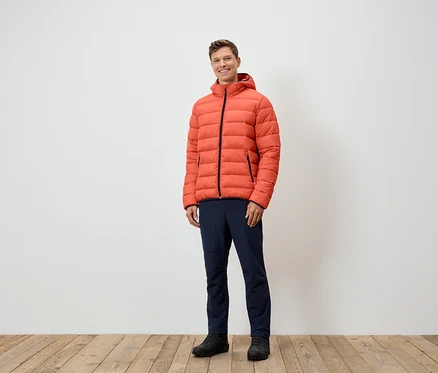 Ein Mann steht und trägt eine orangefarbene Daunenjacke, marineblaue Hosen und schwarze Schuhe.