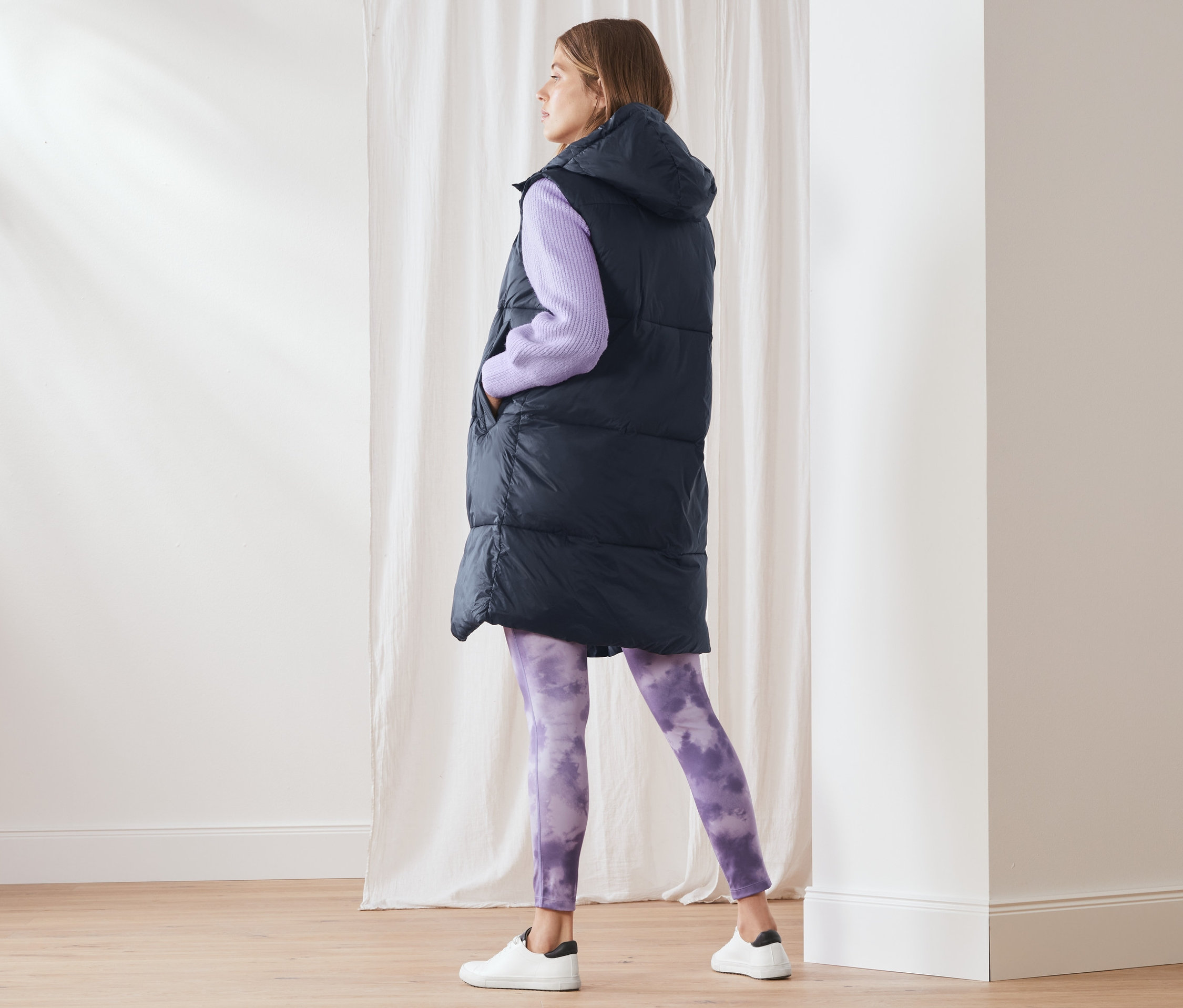 Eine Frau steht und trägt eine lange Steppweste mit Kapuze, einen lila Pullover und lila Leggings.