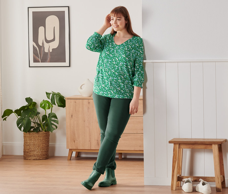 Eine Frau lehnt an der Wand und trägt ein grünes Blusenshirt mit 3/4-Arm und floralem Alloverprint, eine Bengalin-Hose und Regenstiefeletten.