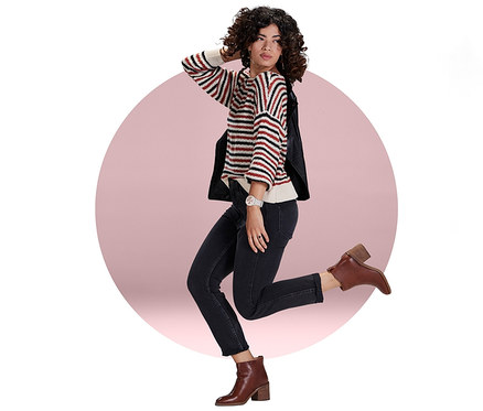 Frau posiert mit einem Bein in der Luft und trägt einen Streifenpullover, Jeans, eine Jacke und braune Stiefel. Sie trägt eine elfenbein-perlgoldfarbene GARMIN Smartwatch »VIVOMOVE® SPORT«.
