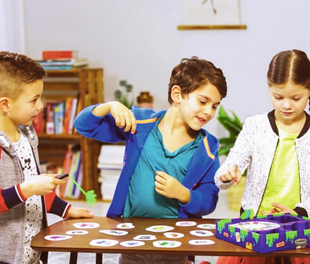 Drei Kinder spielen das Spiel Ravensburger »Monsterstarker Glibberklatsch« auf einem Tisch.
