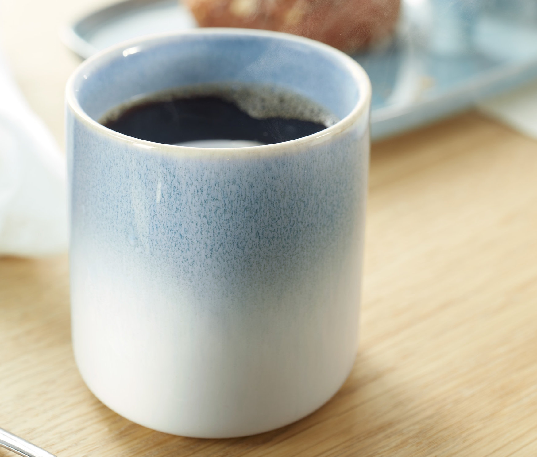 Nahaufnahme eines Kaffeebechers. Im Hintergrund ist ein Teller mit Essen zu sehen.