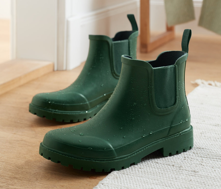 Zwei grüne Regenstiefeletten stehen auf einem Holzboden neben einem weißen Teppich.