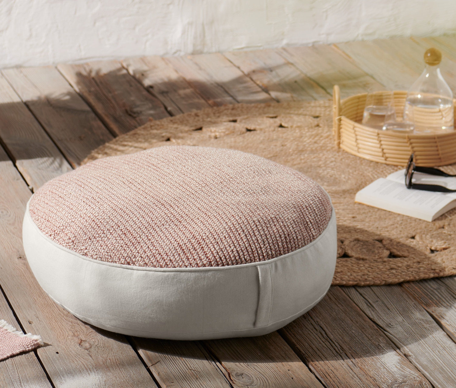 Ein braunes Sitzkissen auf einem Holzboden. Auf einem runden Jute-Teppich steht ein Tablett mit einer Flasche und Gläsern.