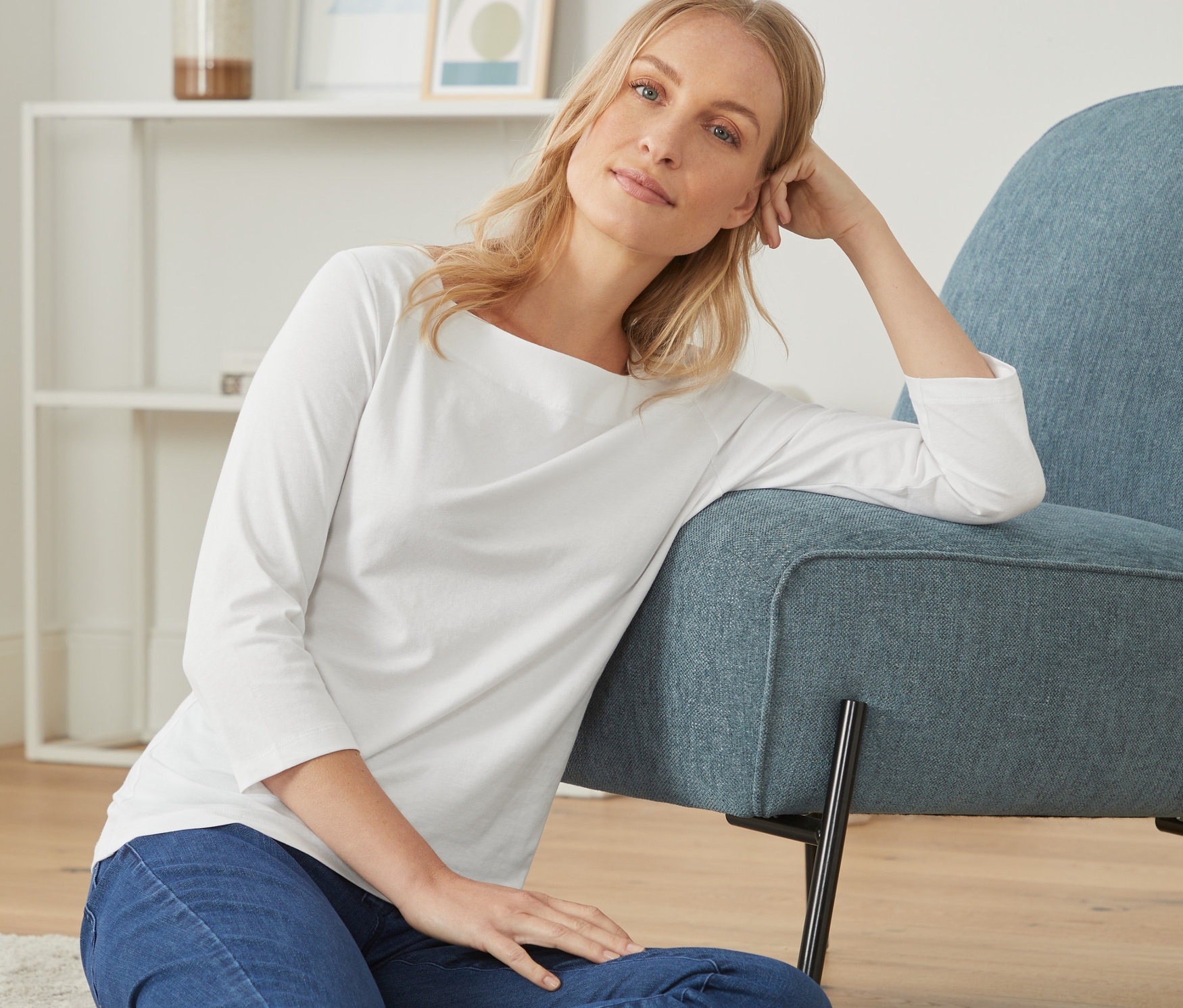 Frau sitzt auf dem Boden und lehnt sich an einen blauen Sessel. Sie trägt ein weißes Shirt mit 3/4-Arm.