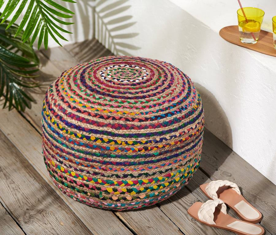 Ein mehrfarbiger Sitzpouf aus Jute steht auf einem Holzboden neben Sandalen.