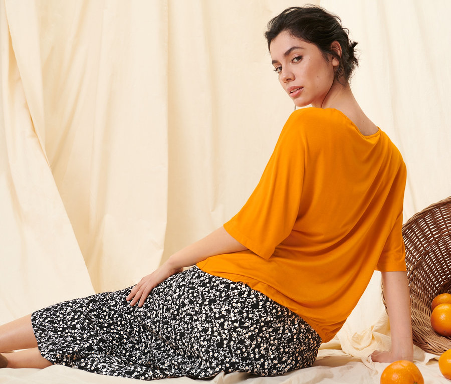 Eine Frau sitzt in einem orangen Blusenshirt und einem floralen Midi-Rock neben einem Korb mit Orangen auf dem Boden.