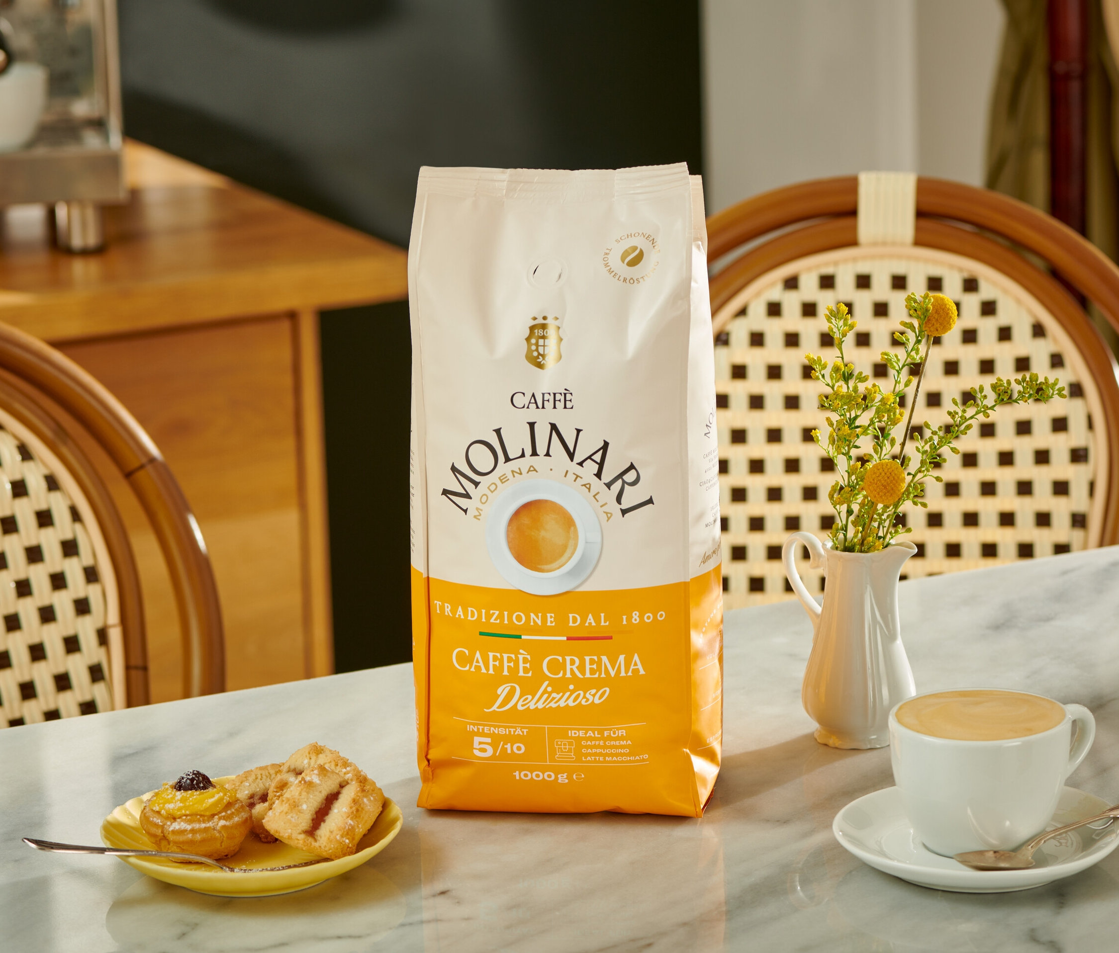 Auf einem Tisch stehen eine Packung Caffè Molinari Caffè Crema Delizioso, Gebäck auf einem gelben Teller, eine Blumenvase und eine Tasse Kaffee.