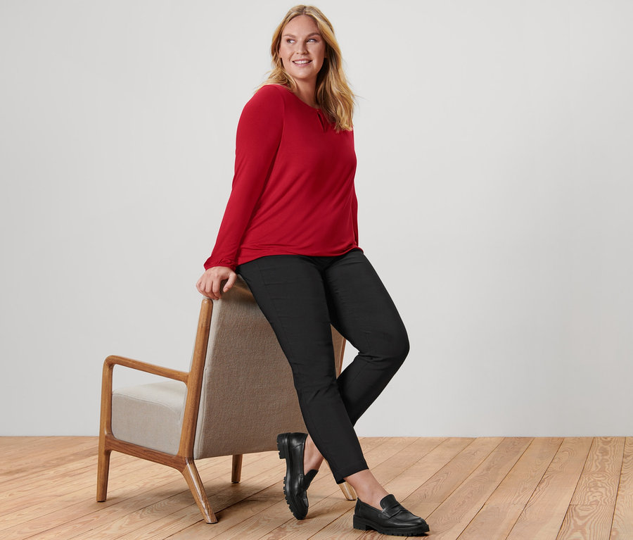 Eine Frau sitzt auf einem Sessel und trägt ein rotes Blusenshirt, eine schwarze Bengalin-Hose und Leder-Loafer.