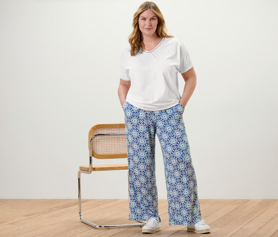 Eine Frau posiert in einem weißen Blusenshirt mit Crochet-Tape und einer Palazzohose mit Alloverprint.