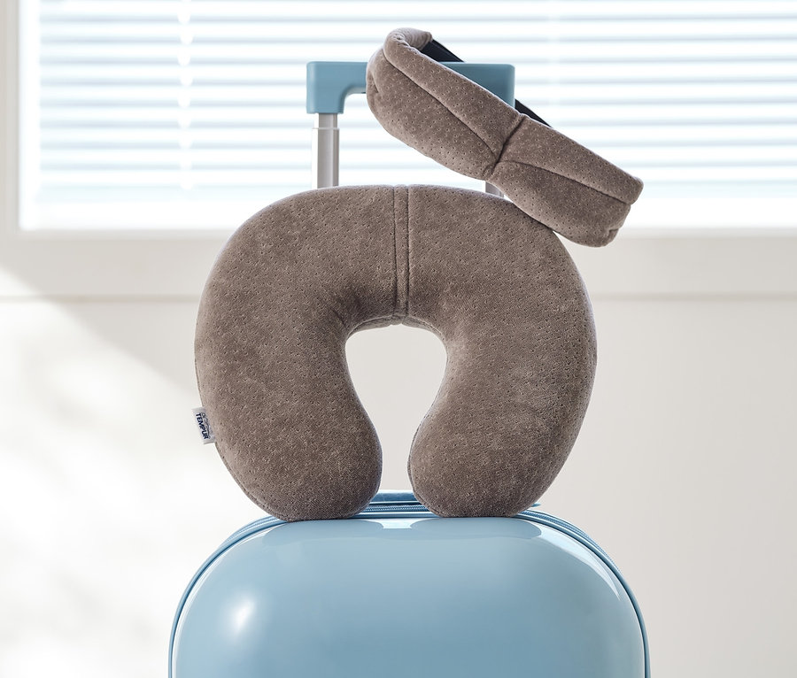 Ein graues TEMPUR® Transitkissen und eine Augenmaske liegen auf einem blauen Koffer vor einem Fenster.