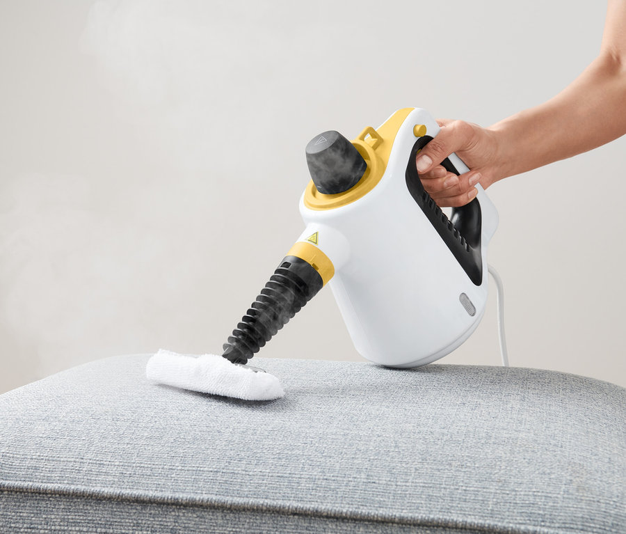 Eine Hand benutzt einen Dampfreiniger auf einem grauen, gepolsterten Hocker.