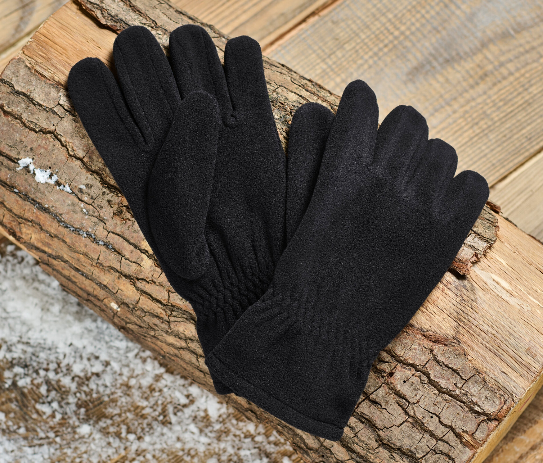 Ein Paar schwarze Microfleece-Handschuhe liegen auf einem Holzscheit vor einem Holzhintergrund.