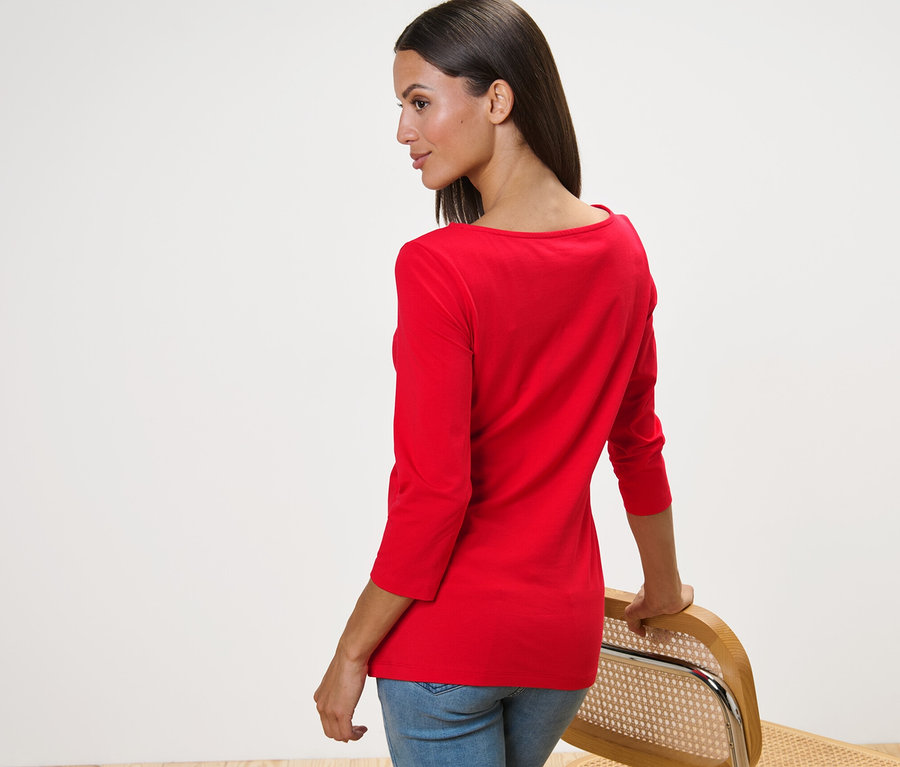 Eine Frau schaut über die Schulter und stützt sich auf einem Stuhl ab, sie trägt ein rotes Shirt mit 3/4-Arm.