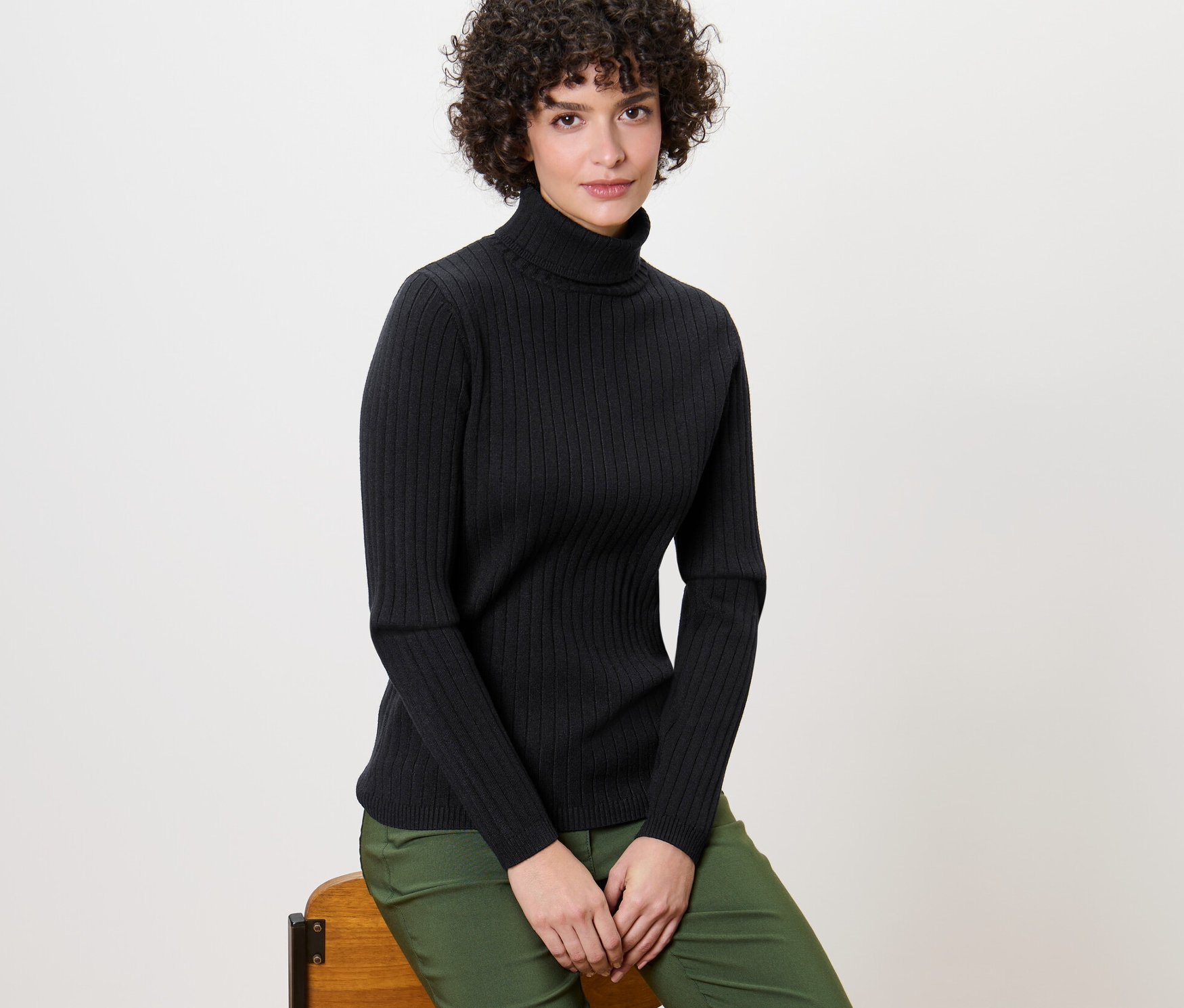 Frau sitzt auf einem Stuhl und trägt einen gerippten schwarzen Rollkragenpullover.