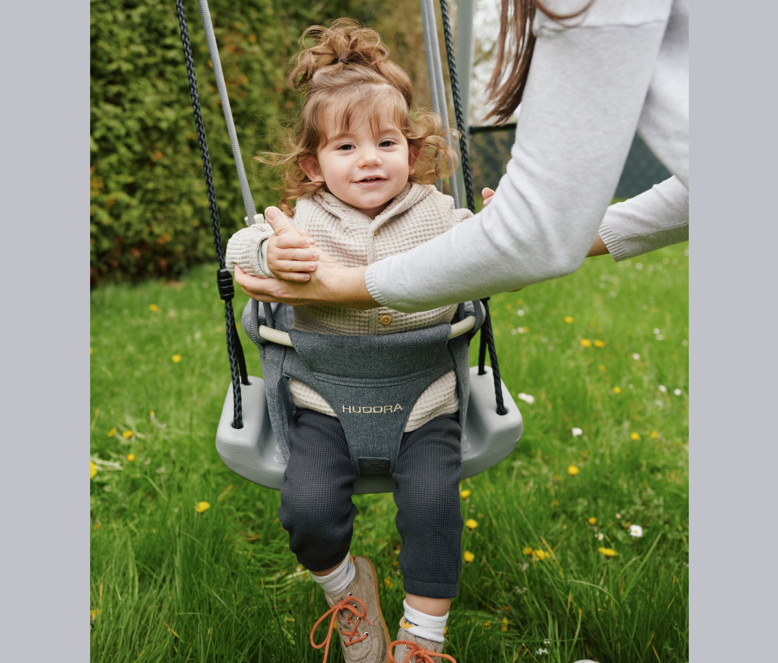 Ein Kleinkind lacht und schaukelt auf einer grauen HUDORA Schaukel, die von einer Frau gehalten wird.