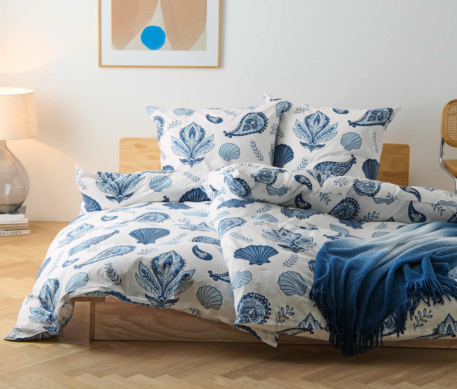 Renforcé-Bettwäsche mit Muschel- und Blumenmuster auf einem Bett. Im Hintergrund eine Lampe und ein abstraktes Bild.