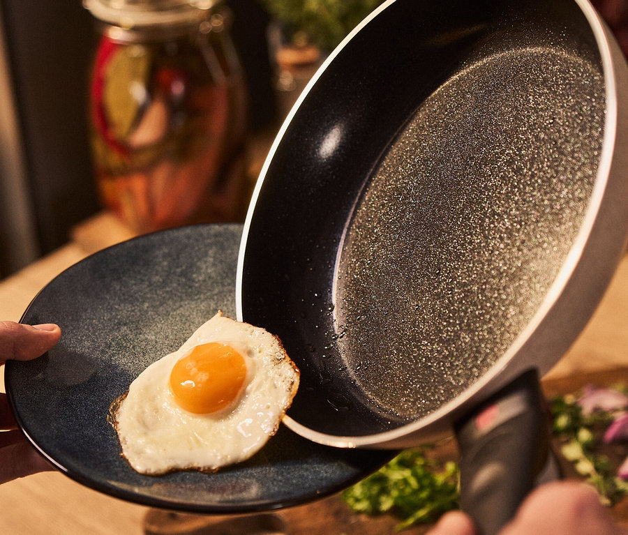 Hand kippt sandfarbene BALLARINI Aluminium-Bratpfanne, 24 cm, um Spiegelei auf blauen Teller gleiten zu lassen.