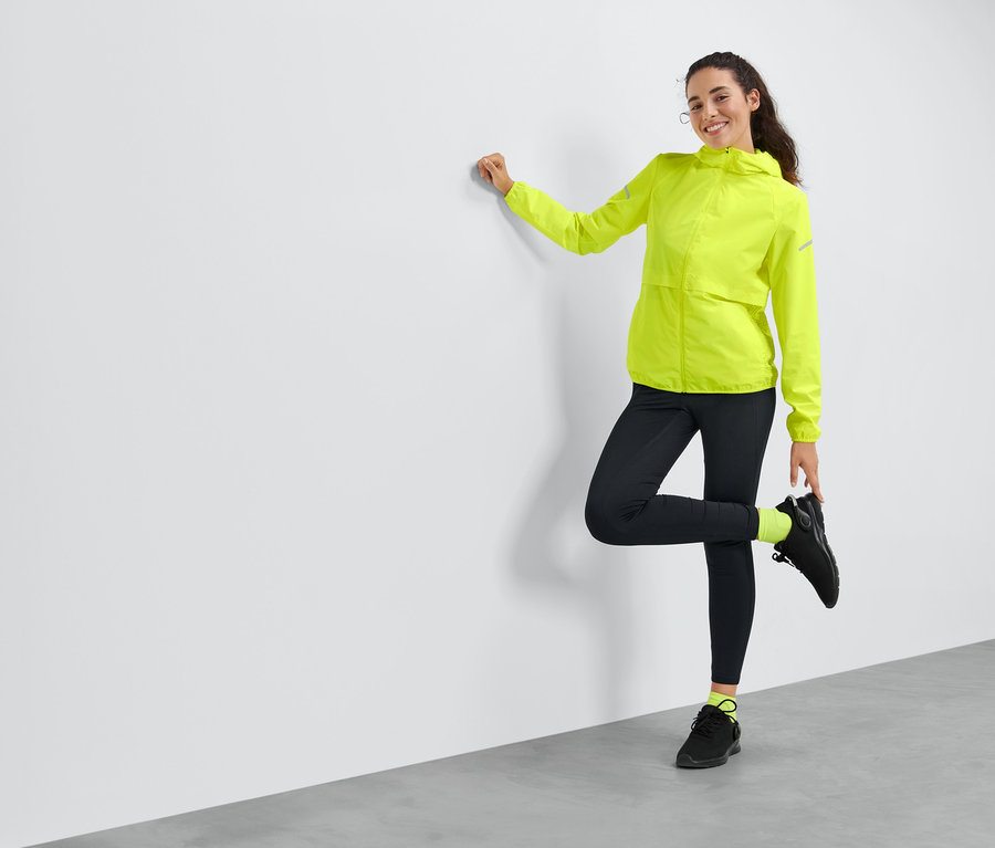 Frau posiert an einer Wand in gelber Windprotection-Laufjacke.