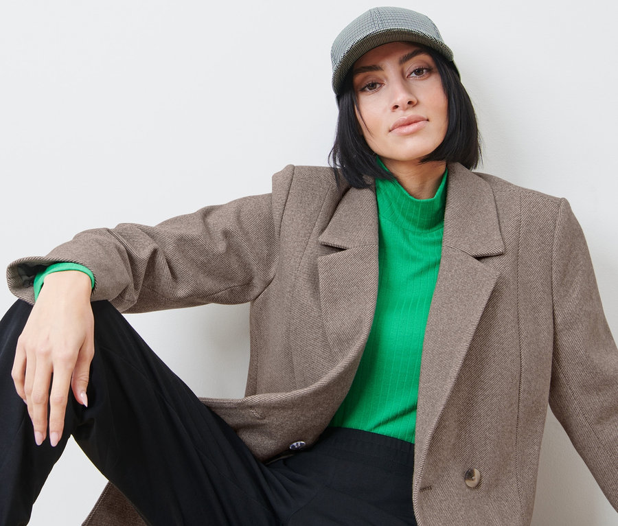 Frau sitzt und trägt einen Blazer, einen grünen Rollkragenpullover und eine Schirmmütze.