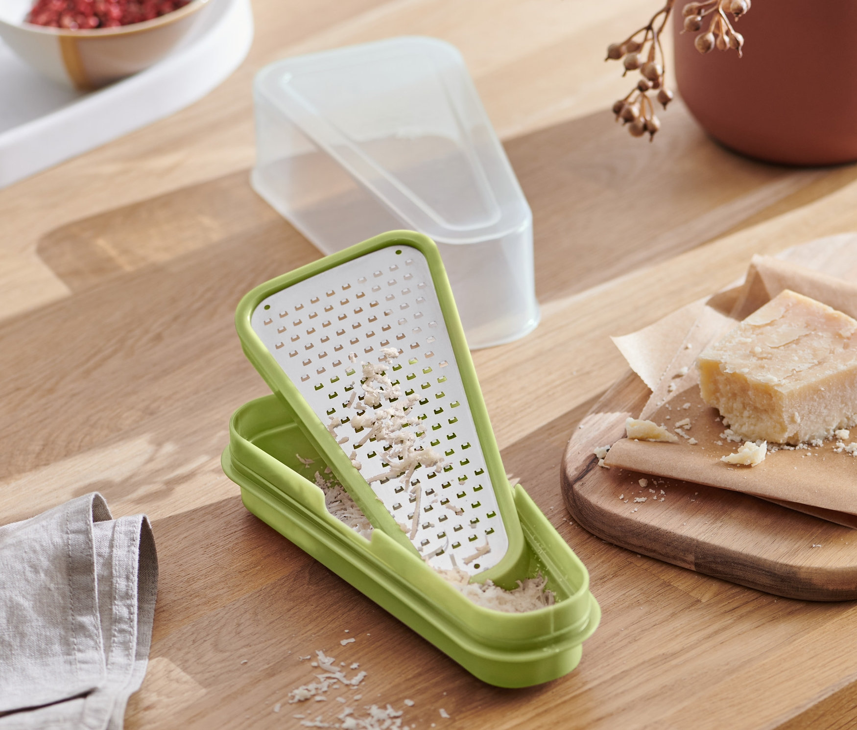 Eine grüne 2-in-1-Parmesanreibe mit geriebenem Käse auf einem Holztisch mit Essen.