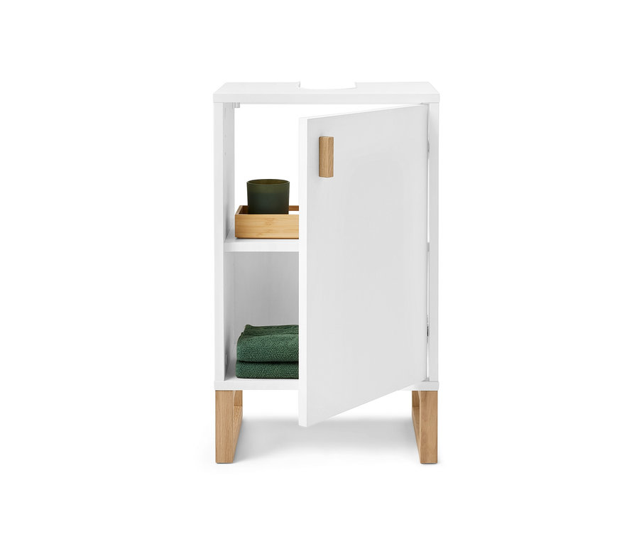 Kleiner weißer Waschbeckenunterschrank mit Tür, die Tür ist offen, innen eine grüne Kerze und grüne Handtücher.