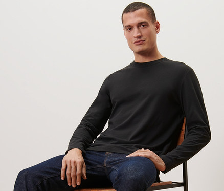 Mann sitzt auf einem Stuhl und trägt ein schwarzes 2 Langarmshirts, Jeans und Turnschuhe.