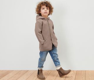 Ein lockiger Junge trägt eine Kinder-Walkjacke mit Merinowolle, Jeans und braune Stiefel.