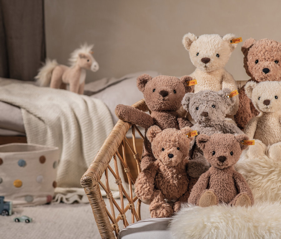 Sieben Stoffteddys in verschiedenen Farben in einem Weidenkorb auf einem Fell. Im Hintergrund ein Bett mit einem Stoffpferd und ein Stoffkorb mit bunten Punkten.
