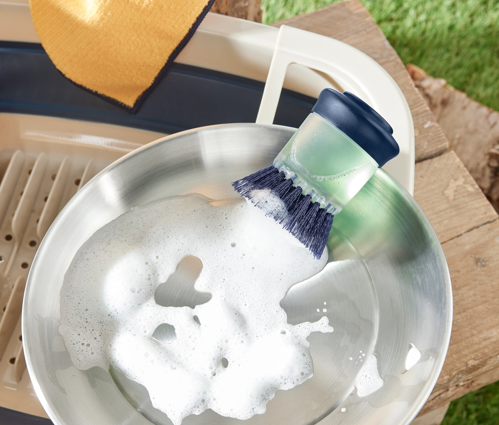 Eine Edelstahlpfanne wird mit einer Spülbürste mit Spender gereinigt. Daneben befinden sich ein Abtropfgestell und ein Lappen.