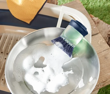 Eine Edelstahlpfanne wird mit einer Spülbürste mit Spender gereinigt. Daneben befinden sich ein Abtropfgestell und ein Lappen.