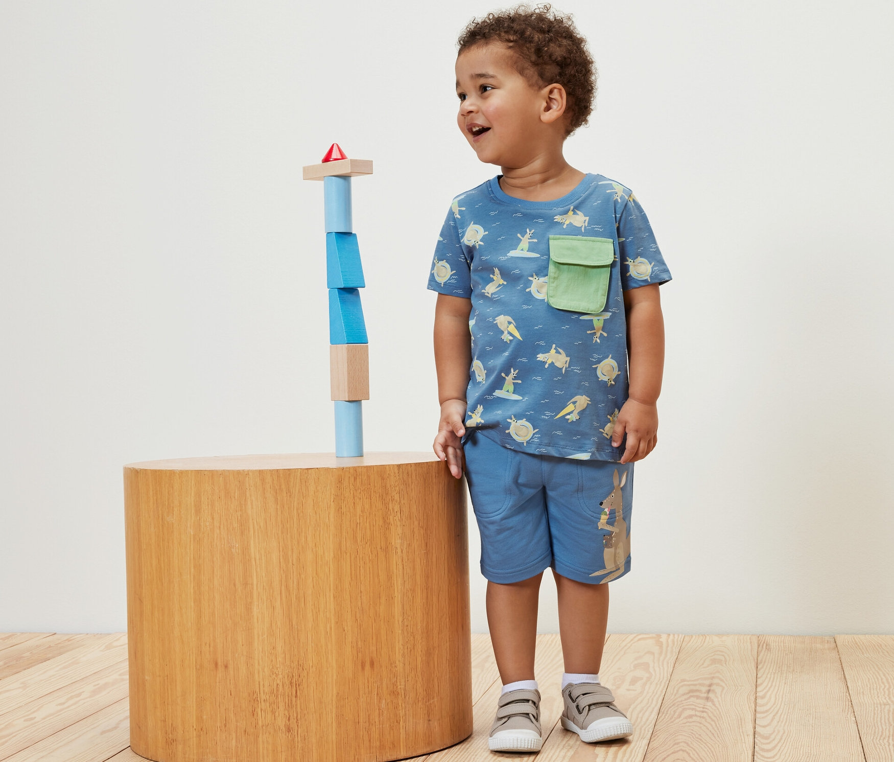 Ein Junge steht neben Holzbausteinen und trägt ein Kinder-T-Shirt.