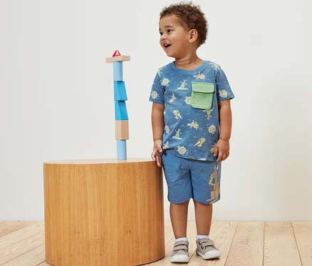 Ein Junge steht neben Holzbausteinen und trägt ein Kinder-T-Shirt.