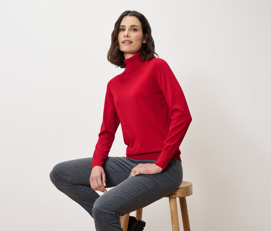 Eine Frau sitzt auf einem Hocker und trägt einen roten Rollkragenpullover und graue Hosen.