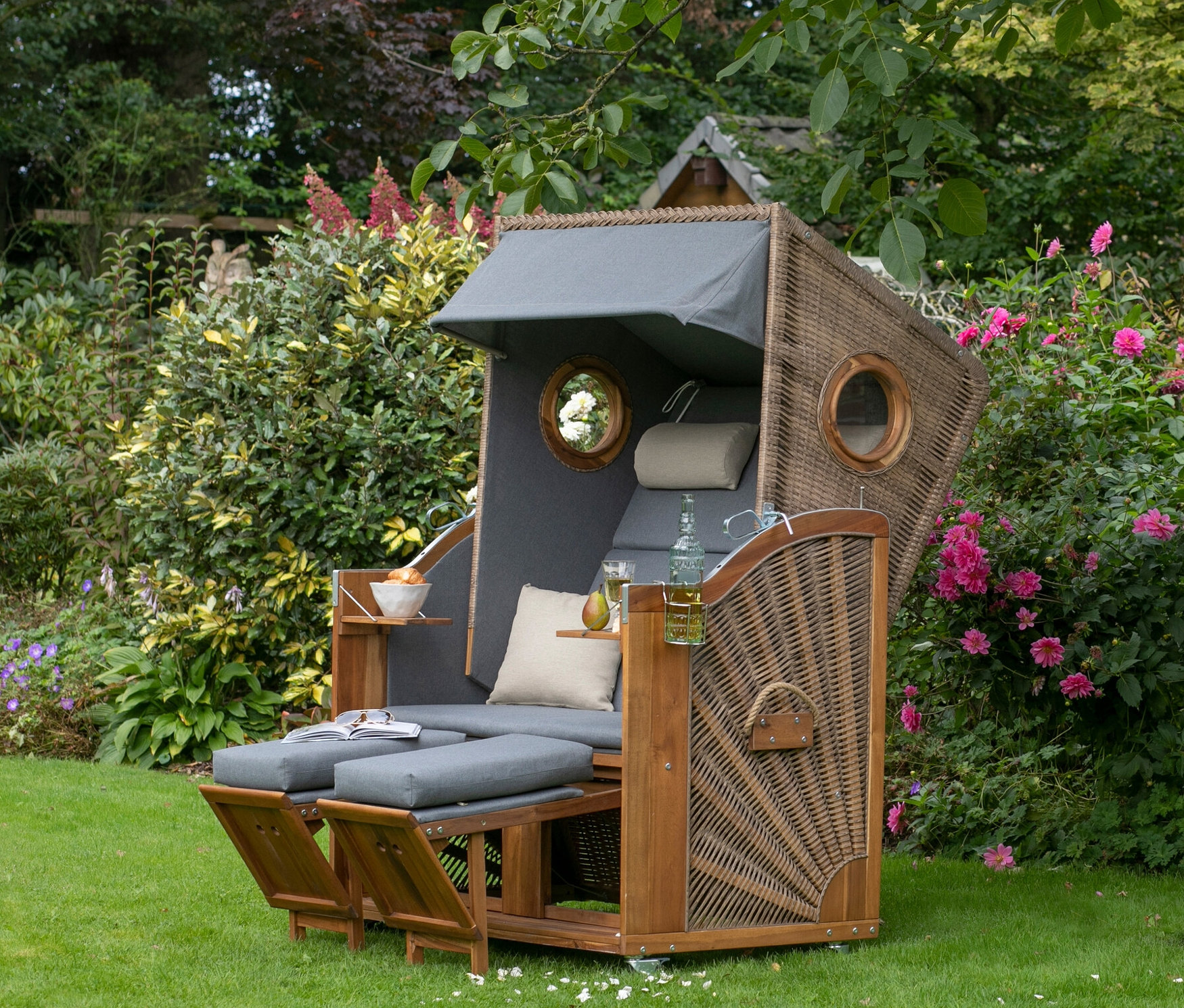 Ein deVries Strandkorb XL aus Korbgeflecht mit grauen Kissen steht auf einer Wiese neben einem Baum.