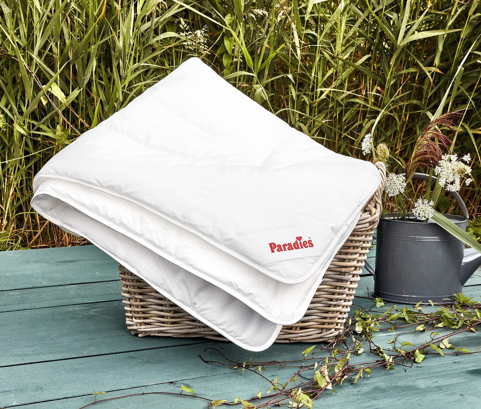 Eine weiße Paradies Sommerdecke »AlsterTraum« liegt gefaltet in einem Weidenkorb neben einer Gießkanne und Grünpflanzen.