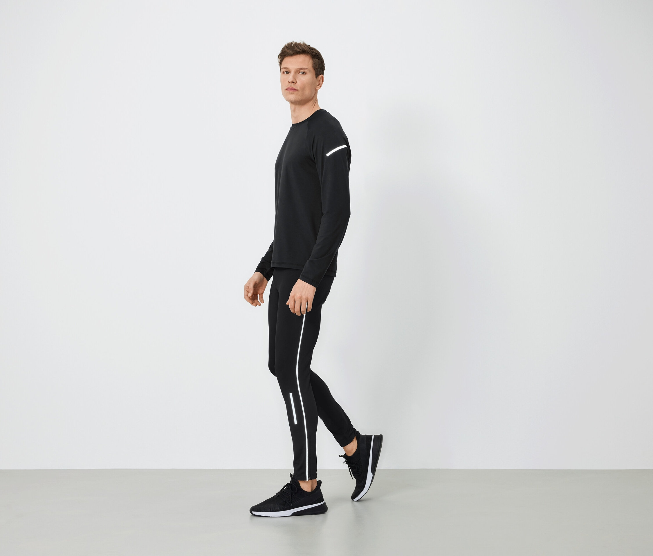 Mann in schwarzer Sportbekleidung mit reflektierenden Details geht auf grauem Boden.