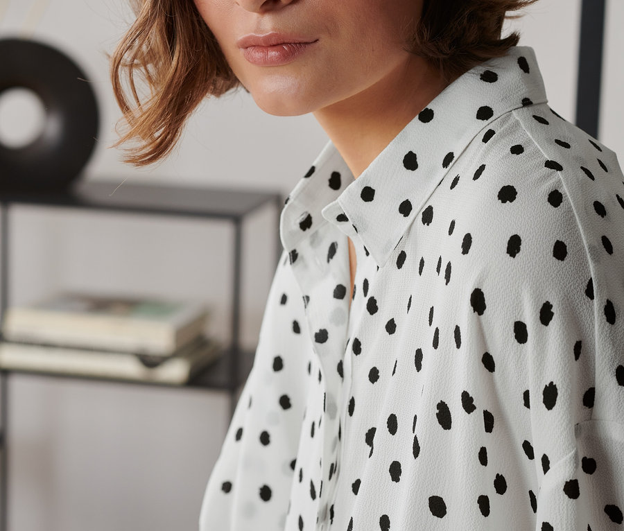 Frau mit braunen Haaren und blauen Augen trägt eine weiße, schwarz gepunktete, kurzärmlige Webbluse.