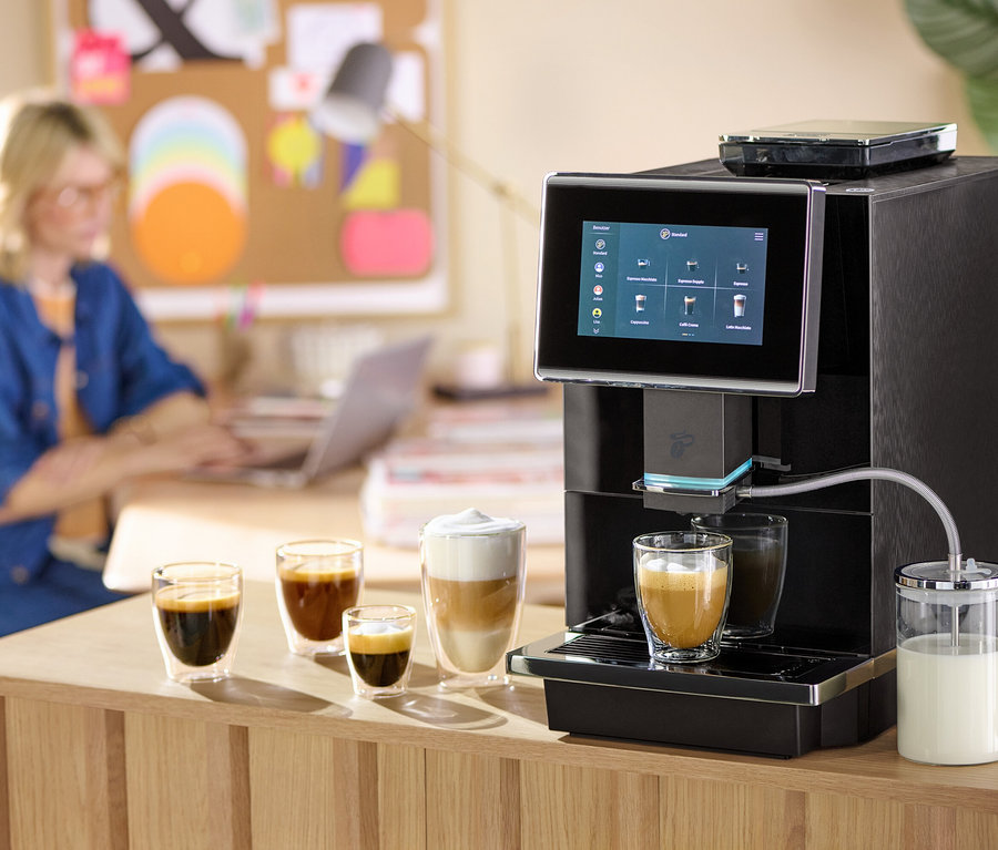 Frau arbeitet im Hintergrund am Laptop, im Vordergrund bereitet eine Kaffeemaschine Kaffee in einem Glas zu.