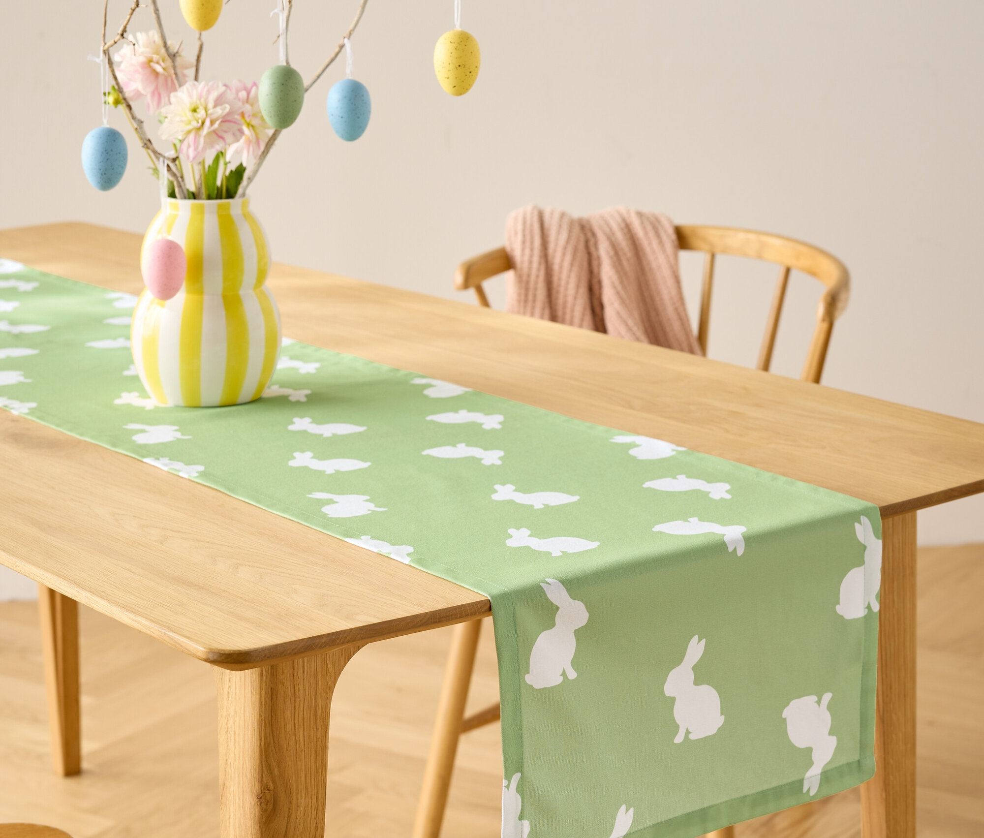 Esstisch mit hellgrünem Tischläufer mit weißen Hasenmotiven, Vase mit Blumen und Ostereier-Dekoration.