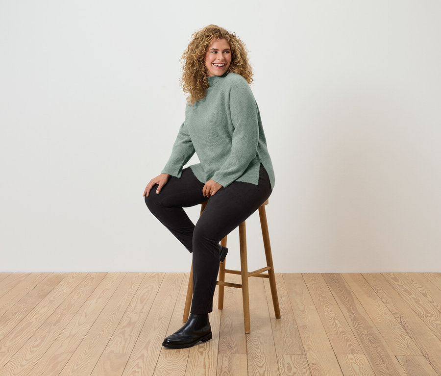 Eine Frau mit lockigem Haar sitzt auf einem Holzhocker und trägt einen grünen Pullover, schwarze Hosen und schwarze Stiefel.