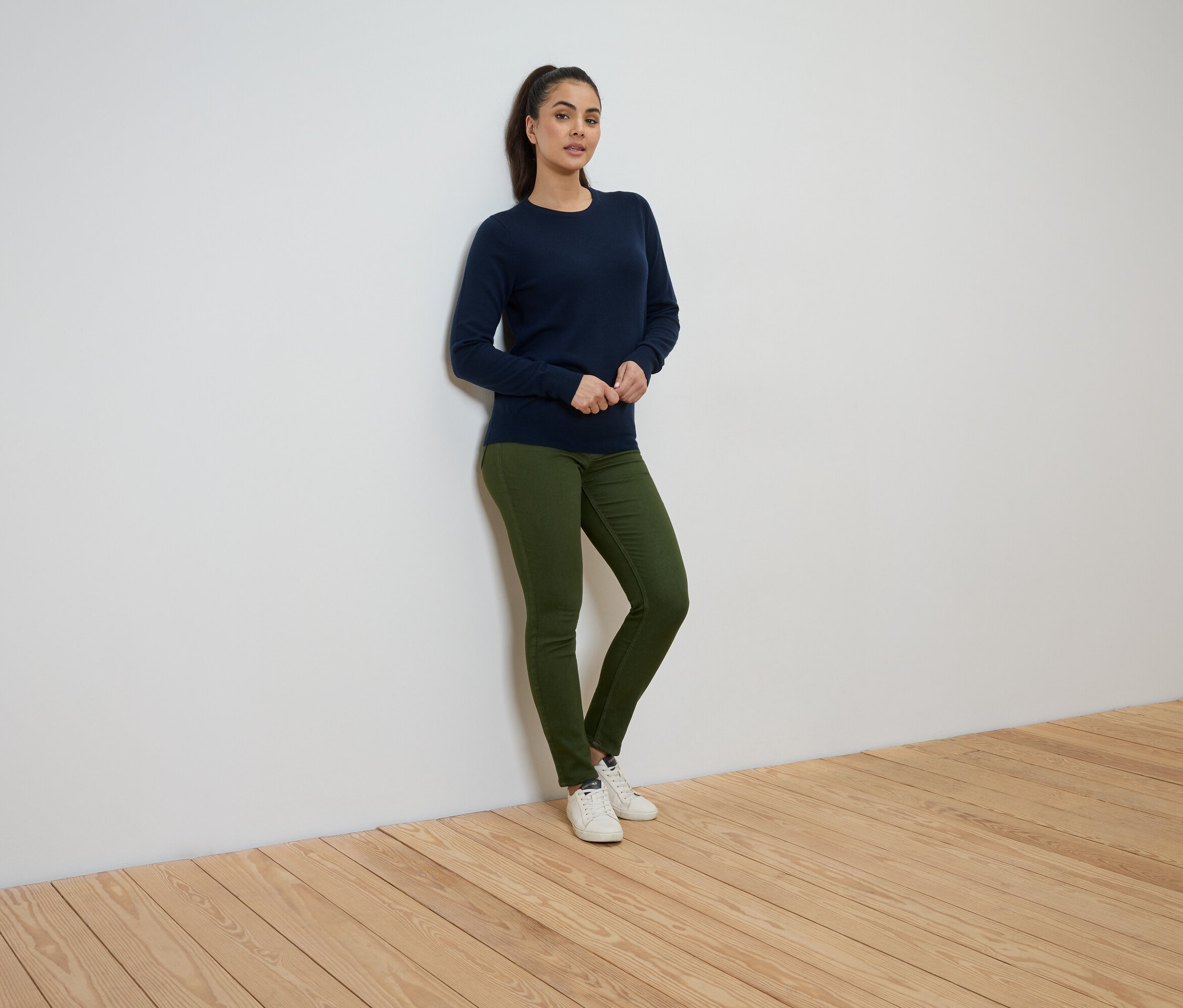 Eine Frau steht an einer Wand und trägt einen marineblauen Pullover, grüne Jeans und weiße Turnschuhe.