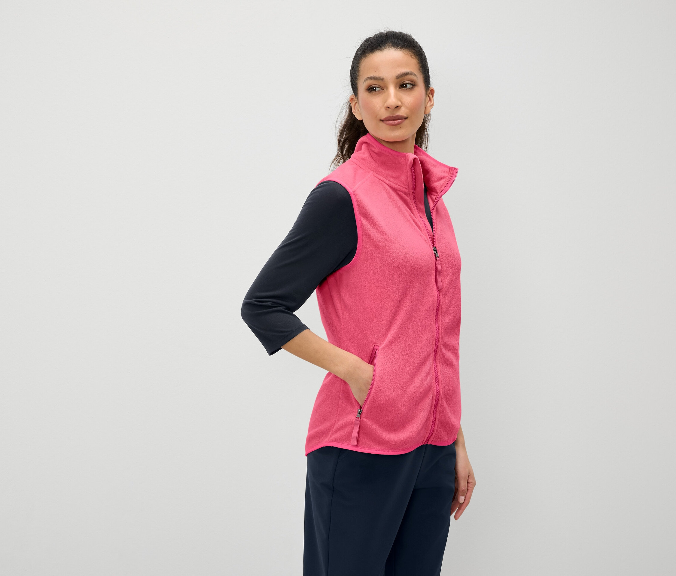 Eine Frau mit braunen Haaren hat ihre Hand in der Tasche einer pinkfarbenen Weste. Sie trägt ein marineblaues Dreiviertelarm-Shirt und marineblaue Hosen.
